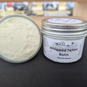 May include: Two small glass jars of whipped tallow balm with silver lids. One jar is open, revealing a creamy, off-white balm with dark specks. The other jar is closed, with a label that reads "Whipped Tallow Balm Vanilla Bean."