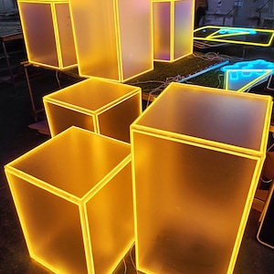 May include: A collection of illuminated, translucent, square and rectangular boxes with bright yellow neon outlines. The boxes are arranged on a surface, with some standing upright and others lying flat. The overall effect is a modern, glowing display.