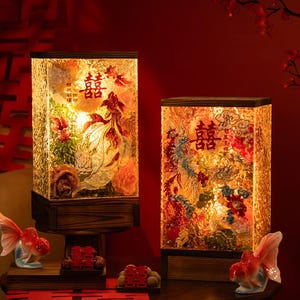 May include: Two illuminated rectangular lamps with wooden bases. The lamps feature intricate designs of fish and flowers, with Chinese characters. Decorative goldfish figurines and small red boxes are in the foreground.