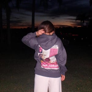 May include: A dark grey hoodie with a graphic print on the back, featuring text and images in pink, white, and black. The person is wearing white trousers. The background is a dark evening scene with palm trees.