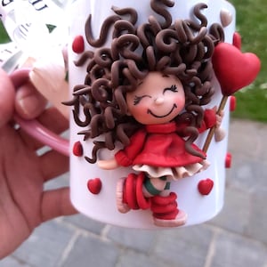 May include: White ceramic mug with a whimsical design featuring a smiling figure in a red dress holding a heart-shaped balloon. The mug is adorned with red hearts and brown curly hair. A pink handle and a ribbon are visible.