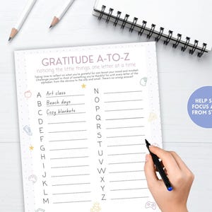 May include: A gratitude journal with the title "Gratitude A-to-Z" and the text "noticing the little things, one letter at a time." The journal has a pink dotted border and includes prompts for each letter of the alphabet. A hand is holding a black pen.
