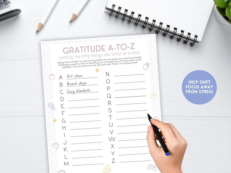 May include: A gratitude journal with the title "Gratitude A-to-Z" and the text "noticing the little things, one letter at a time." The journal has a pink dotted border and includes prompts for each letter of the alphabet. A hand is holding a black pen.