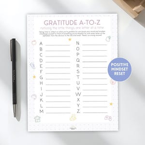 May include: A gratitude journal with the title "GRATITUDE A-TO-Z" and the text "noticing the little things one letter at a time." The journal has a list of the alphabet with lines to write on. A black pen is on the left.