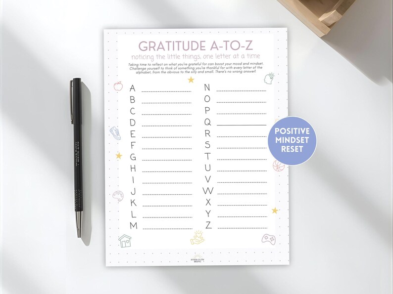 May include: A gratitude journal with the title "GRATITUDE A-TO-Z" and the text "noticing the little things one letter at a time." The journal has a list of the alphabet with lines to write on. A black pen is on the left.