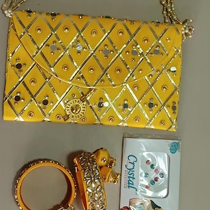 May include: Yellow clutch with a beaded strap, adorned with gold accents and small mirrors. Includes matching yellow bangles and a bracelet with floral mirror details. A package of crystal embellishments is also present.