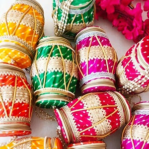 May include: A collection of colorful, decorative drums. The drums are cylindrical with a patterned fabric in shades of yellow, orange, green, and pink. Each drum is adorned with gold trim, white lace, and pearl accents, creating a festive appearance.