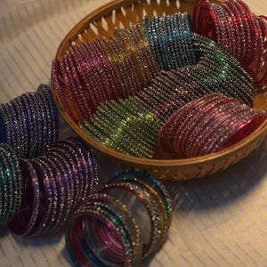 Könnte beinhalten: Eine Sammlung bunter Armreifen in einem geflochtenen Korb. Die Armreifen sind in den Farbtönen Rot, Grün, Blau und Gold gehalten und haben ein schimmerndes, juwelenbesetztes Aussehen. Einige Armreifen liegen außerhalb des Korbs.