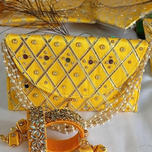Puede incluir: Un bolso de mano amarillo con un patrón geométrico, adornado con detalles dorados y pequeñas gemas. También se ven un brazalete y pendientes a juego, junto con un collar de perlas. El conjunto es probablemente para ocasiones especiales.