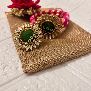 May include: Gold-toned earrings with green centres and clear stone accents, alongside a matching bracelet and a red flower. The jewellery sits on a gold textured pouch.