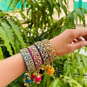 Puede incluir: Un conjunto de pulseras coloridas con diseños florales y adornos colgantes. Las pulseras son en tonos turquesa, rosa, negro y amarillo, adornadas con detalles plateados. El fondo presenta follaje verde.