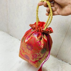 Può includere: Una borsa rossa e dorata con coulisse e manico in corda dorata. La borsa presenta un motivo floreale ornato in oro e verde. Il cordoncino è rosa intenso e il nappa è rosso.
