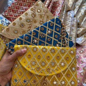 Könnte beinhalten: Eine Kollektion rechteckiger Umschlag-Clutches mit einem rautenförmigen Goldbesatz. Jede Tasche ist mit kleinen runden Spiegeln und metallischen Blumenakzenten verziert. Die Taschen sind in den Farben Gelb, Blau und Beige erhältlich.