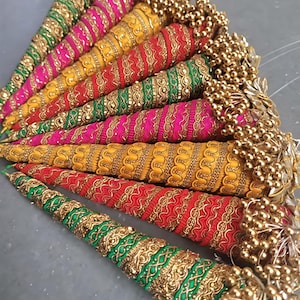 May include: A collection of colorful, cone-shaped decorative items. Each cone is wrapped in vibrant fabric, including green, pink, yellow, and red, and adorned with gold-colored embellishments and beaded tassels. The items are arranged in a fan-like pattern.