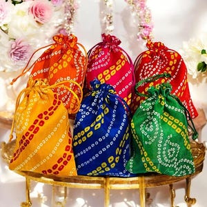 Op de afbeelding: Een verzameling kleurrijke trekkoordzakken in oranje, geel, rood, roze, blauw en groen. Elke tas heeft een uniek patroon van kleine, zich herhalende vormen en een trekkoordsluiting. De tassen zijn gerangschikt op een goudkleurig, sierlijk dienblad.