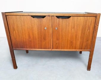 Può includere: Un mobile in legno in stile moderno della metà del secolo con due ante e un piano scuro. Il mobile ha una finitura marrone caldo, maniglie incassate nere e pomelli argentati. Le gambe sono angolate.