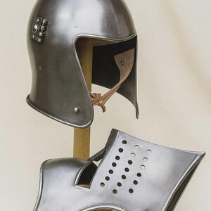 May include: A medieval steel helmet and gorget, displayed on a wooden stand. The helmet has a rounded top and a face opening. The gorget, with a series of small holes, is designed to protect the neck and shoulders. The metal has a dark, reflective finish.