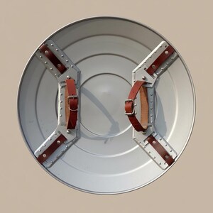 May include: A circular, silver-coloured shield with riveted metal supports and brown leather straps. The shield has a slightly concave centre and is designed for defence. The straps are buckled for a secure grip.