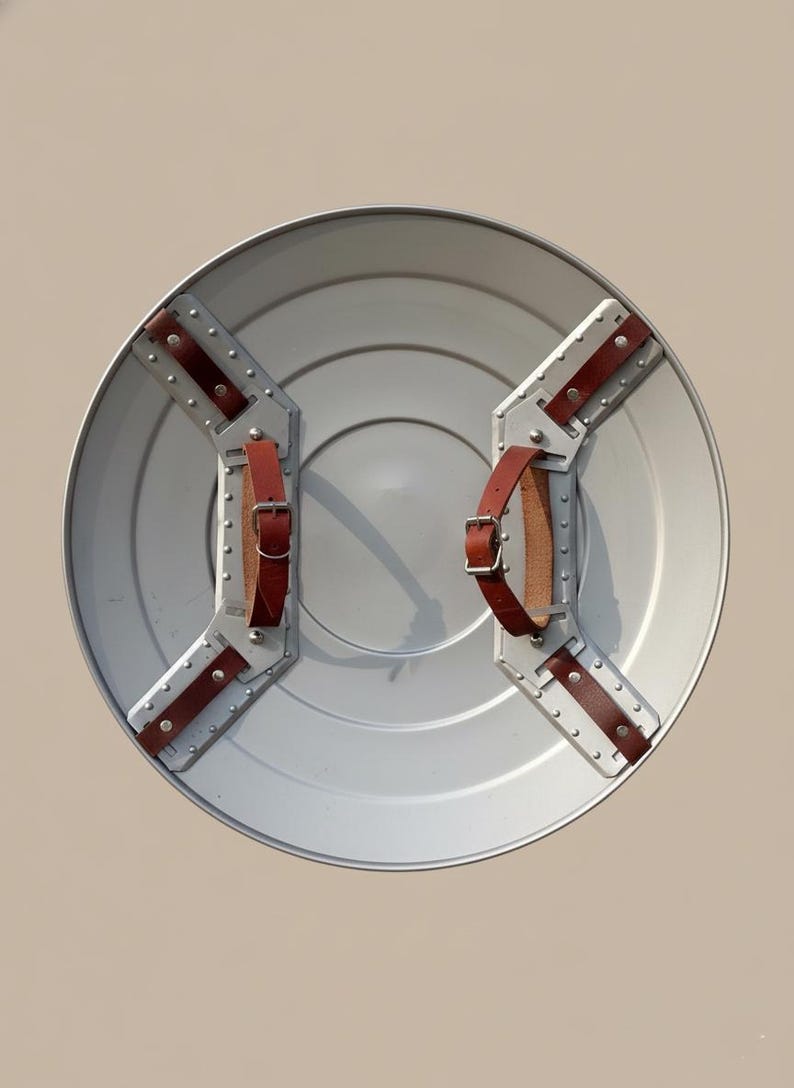 May include: A circular, silver-coloured shield with riveted metal supports and brown leather straps. The shield has a slightly concave centre and is designed for defence. The straps are buckled for a secure grip.