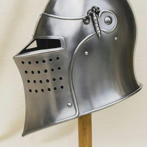 May include: A polished silver-colored medieval helmet with a rounded top and a visor featuring small ventilation holes. The helmet is displayed on a wooden stand, showcasing its historical design and craftsmanship. A chain is attached to the side.