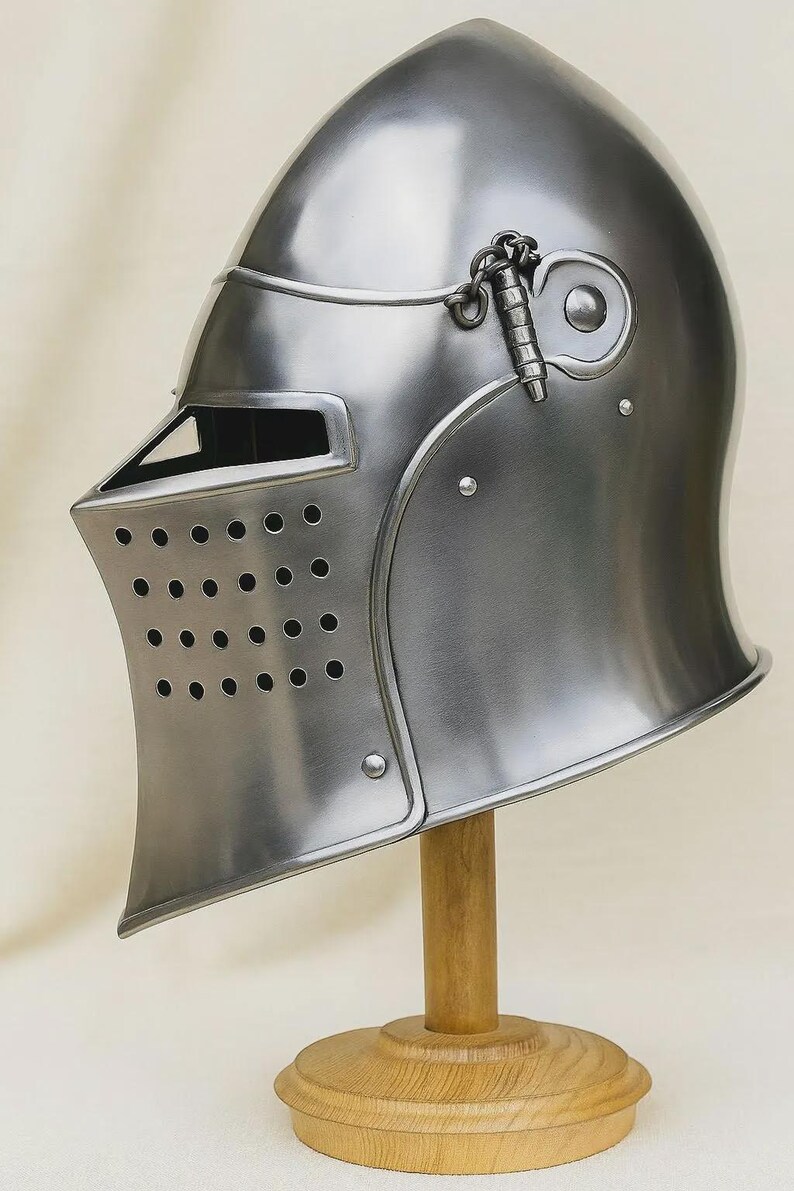 May include: A polished silver-colored medieval helmet with a rounded top and a visor featuring small ventilation holes. The helmet is displayed on a wooden stand, showcasing its historical design and craftsmanship. A chain is attached to the side.