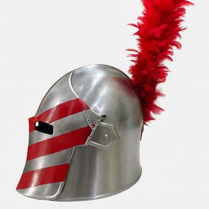 May include: A silver-colored knight's helmet with red stripes on the visor and a chain detail. A large plume of red feathers extends from the top of the helmet. The helmet is likely made of metal and is designed for protection.