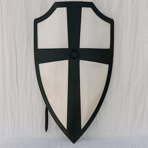 May include: A shield featuring a black cross on a white background, framed by a black border. The shield has a pointed base and a curved top. A black strap is attached.