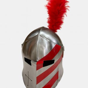 May include: A polished metal knight's helmet with red and white stripes and a red plume. The helmet has a cross design on the front and eye slits. A chain is attached to the side of the helmet.