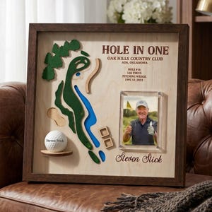 May include: A wooden shadow box display featuring a 3D map of a golf course hole, a white golf ball on a small shelf, and a photograph of a person. The text reads HOLE IN ONE, OAK HILLS COUNTRY CLUB, ADA, OKLAHOMA, HOLE #16, LAK PSKUS, PITCHING WEDGE, 199T 12, 2023, and Steven Stick.