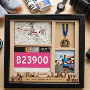 May include: A black framed shadow box display featuring a marathon race bib with number B23900, a topographical map, a gold medal with a blue and yellow ribbon, and a photograph of a runner. The frame includes the text JOHN, BOSTON MARATHON 2024, 2:49:21.