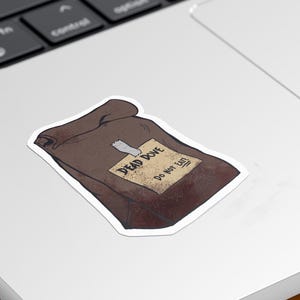 May include: A brown sticker in the shape of a bag, with a white border, is affixed to a laptop. The sticker has a label that reads "DEAD DOVE Do not eat". The laptop is on a yellow surface.