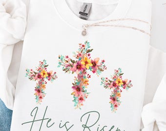 May include: White sweatshirt featuring three floral crosses in pink, yellow, and green. The phrase "He is Risen" is written in green script below the crosses. A silver necklace with a small pendant is draped across the sweatshirt.