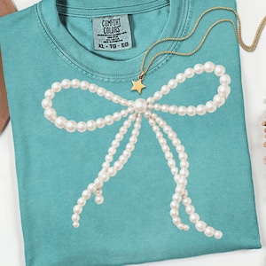 May include: A turquoise Comfort Colors t-shirt featuring a pearl bow design. A gold necklace with a star pendant rests on the shirt. The shirt has a crew neck and short sleeves. The tag reads XL - TG - EG.