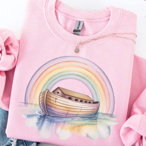 Puede incluir: Sudadera rosa claro con una ilustraci&oacute;n en acuarela del Arca de No&eacute; bajo un arco&iacute;ris. El dise&ntilde;o muestra un arca de madera sobre el agua con un arco&iacute;ris encima. Un collar dorado con un colgante de sol descansa sobre la sudadera.