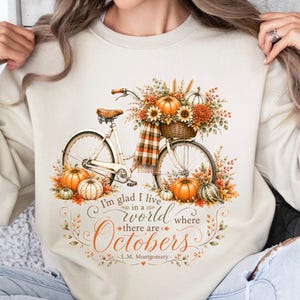 May include: A beige crewneck sweatshirt featuring a graphic of a vintage bicycle with a basket filled with sunflowers and pumpkins. The text reads: I'm glad I live in a world where there are Octobers - L.M. Montgomery, surrounded by autumn leaves.
