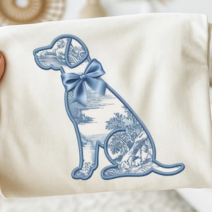 May include: A cream-colored fabric item featuring a blue outline of a dog with a bow tie. The dog's interior is filled with a blue and white toile pattern depicting a landscape scene. The bow tie is a solid blue color.
