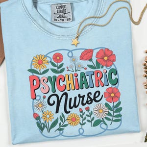 May include: Light blue t-shirt with the words "PSYCHIATRIC Nurse" in a colorful, retro font, surrounded by floral illustrations. A gold star pendant necklace rests on the shirt. The shirt is a Comfort Colors XL-TG-EG.