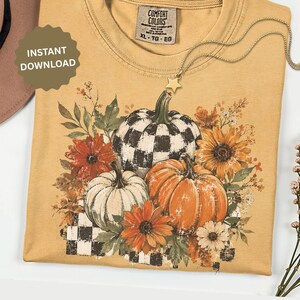 May include: A mustard yellow t-shirt featuring a graphic print of three pumpkins, including one with a black and white buffalo check pattern, surrounded by sunflowers, autumn leaves, and small white flowers, with a star-shaped necklace charm.