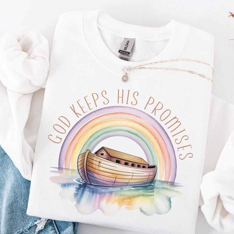 Puede incluir: Sudadera blanca de cuello redondo con el texto "GOD KEEPS HIS PROMISES" sobre una ilustraci&oacute;n de acuarela del Arca de No&eacute; y un arco&iacute;ris. Un collar plateado con un colgante de sol est&aacute; sobre la sudadera.