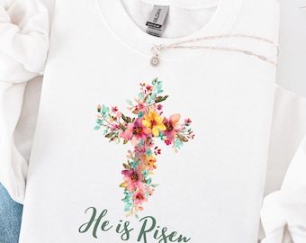 May include: White sweatshirt featuring a floral cross design with pink, yellow, and blue flowers. The words "He is Risen" are printed in green script below the cross. A silver necklace with a small pendant is draped on the sweatshirt.