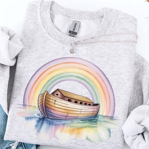 Puede incluir: Una sudadera gris claro con una ilustraci&oacute;n en acuarela del Arca de No&eacute; bajo un arco&iacute;ris. El arca est&aacute; representada en tonos marrones y amarillos, con un arco&iacute;ris arqueado encima. Un collar plateado con un peque&ntilde;o colgante est&aacute; sobre la sudadera.