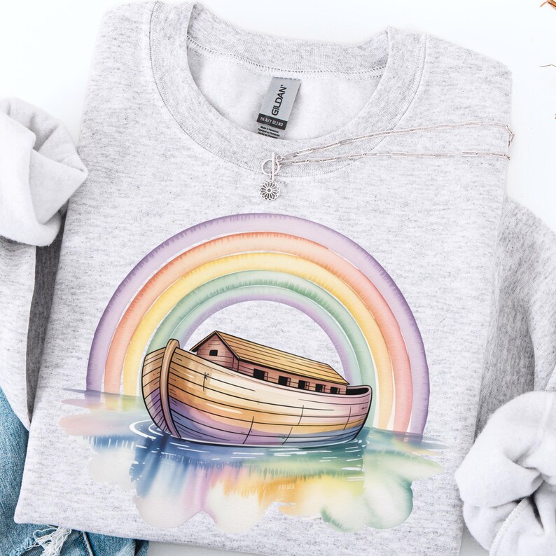 Puede incluir: Una sudadera gris claro con una ilustraci&oacute;n en acuarela del Arca de No&eacute; bajo un arco&iacute;ris. El arca est&aacute; representada en tonos marrones y amarillos, con un arco&iacute;ris arqueado encima. Un collar plateado con un peque&ntilde;o colgante est&aacute; sobre la sudadera.