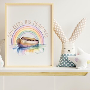 Puede incluir: Una impresi&oacute;n en acuarela enmarcada con un arco&iacute;ris, el Arca de No&eacute; y el texto "GOD KEEPS HIS PROMISES" en un dise&ntilde;o circular. Un conejito de peluche con orejas de lunares y una almohada en forma de estrella est&aacute;n en un estante blanco.