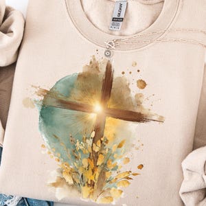 May include: A beige sweatshirt with a watercolor design featuring a brown cross, a teal circle, and yellow and brown floral accents. The design has a bright, sun-like burst of light at the center of the cross. A silver necklace is visible at the neckline.