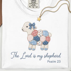 May include: White t-shirt with a patchwork lamb design in red, white, and blue patterns, a blue bow, and the text "The Lord is my shepherd, Psalm 23." A gold star pendant necklace is draped across the shirt.
