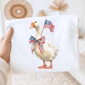 May include: Watercolor illustration of a white goose adorned with a patriotic bow and holding a small American flag. The goose has orange feet and beak. The image is printed on a white fabric.