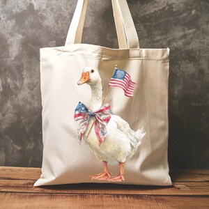 May include: Cream-colored tote bag featuring a watercolor illustration of a white goose adorned with a patriotic bow and holding a small American flag. The bag has sturdy handles and is perfect for carrying groceries or personal items.