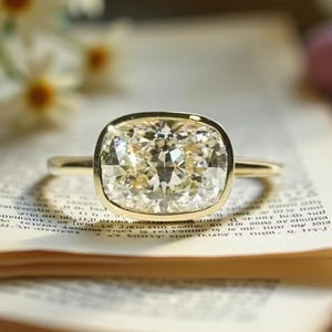 Könnte beinhalten: Ein goldener Ring mit einem großen Diamanten im Kissenschliff. Der Diamant ist in einer Zargenfassung gefasst und liegt auf einem offenen Buch. Der Ring hat eine hellgelbe Farbe und ist vor einem verschwommenen Hintergrund aus Blumen platziert.