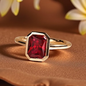 Pode incluir: Um anel de ouro com uma pedra preciosa retangular vermelha escura. A pedra preciosa é engastada em uma moldura de ouro, e a banda é de ouro polido. O anel é exibido em uma superfície marrom.