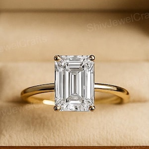May include: A gold engagement ring with a large, rectangular, emerald-cut diamond is displayed inside a wooden ring box. The ring box is open, revealing the ring's setting and the diamond's facets. The box is made of dark wood.